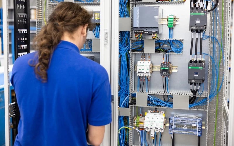 A wireman wearing a blue polo shirt working on a large electrical control cabinet assembly conisting of circuit breakers, relays, fuses and wiring.