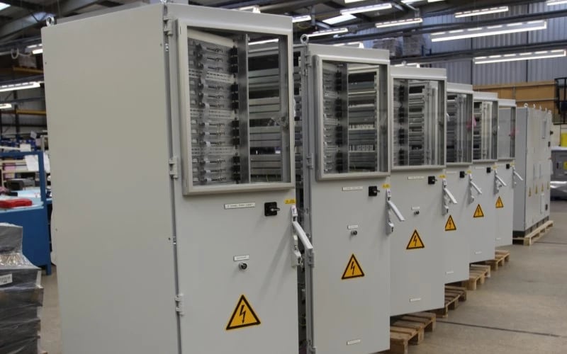 A row of ten large electrical control cabinets secured to wooden pallets with the front doors open waiting for final inspection on a manufacturing shop floor.