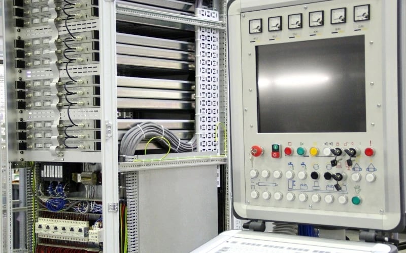 The inside of a large multi-rack electrical control cabinet alongside a human machine interface system which includes a touchscreen display and many electrical buttons.