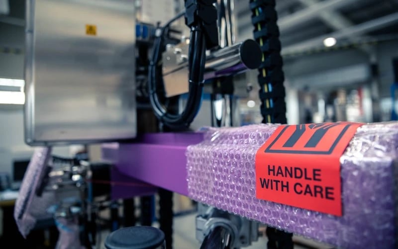 Eine grosse elektromechanische Druck- und Applikationsmaschine mit einem leuchtend violetten Metallarm wartet auf die endgültige Verpackung und den Versand mit einem Griff, an dem ein Pflegeetikett befestigt ist.