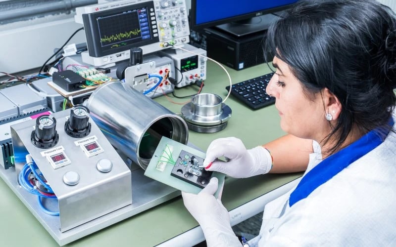 A female production operator wearing white gloves sitting at a bench in front of a custom piece of electronic manufacturing test equipment.  