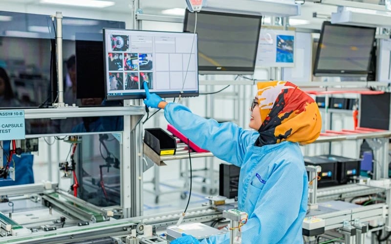 Eine weibliche Produktionsmitarbeiterin mit blauer ESD-Jacke und buntem Kopftuch zeigt auf eine Reihe von Arbeitsanweisungen auf einem elektronischen Bildschirm.