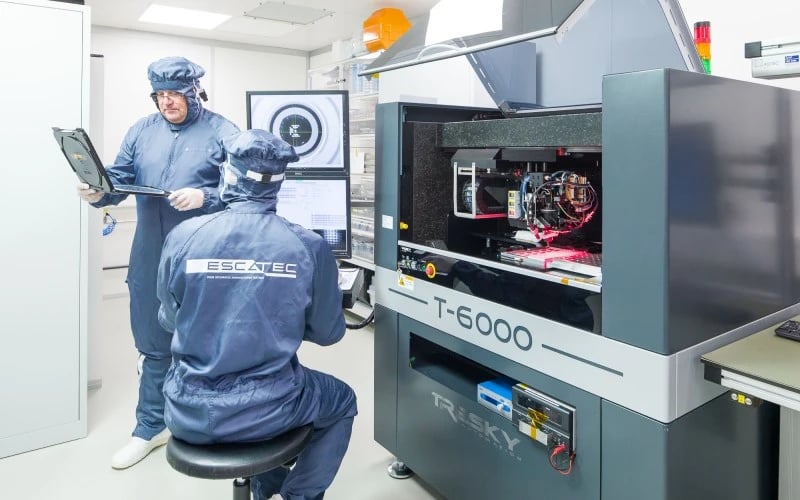 Two electronics manufacturing operatives wearing blue overalls in a clean room environment. One is standing up holding a tray of chip wavers and one is sitting in front of a Tresky T-6000 Die Bonder machine.  