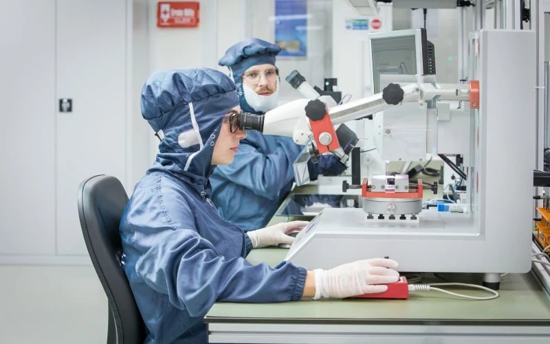 Two electronics manufacturing operatives wearing blue overalls in a clean room environment. One operator is inspecting an electronic microchip under a high power microscope. 