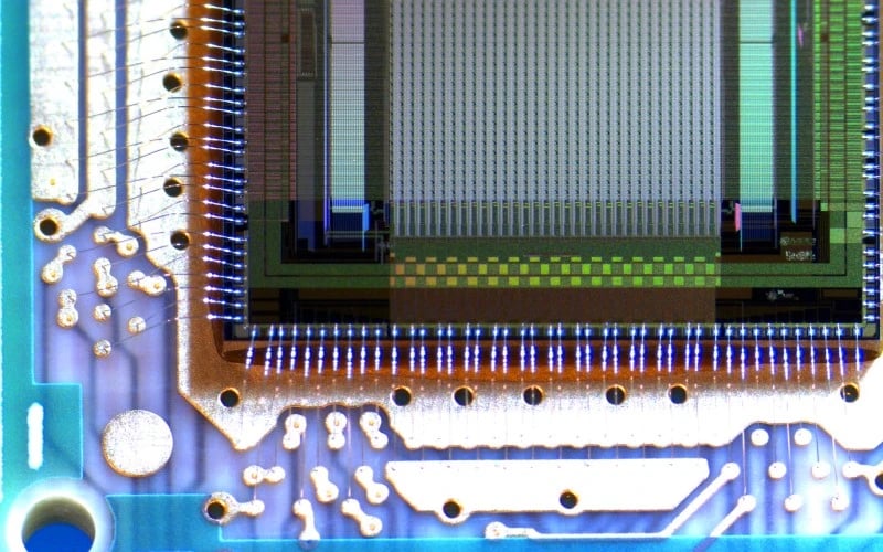 The inside of electronic semiconductor chip under high magnification showing exposed bare wires and the silicon die in green and blue lighting. 