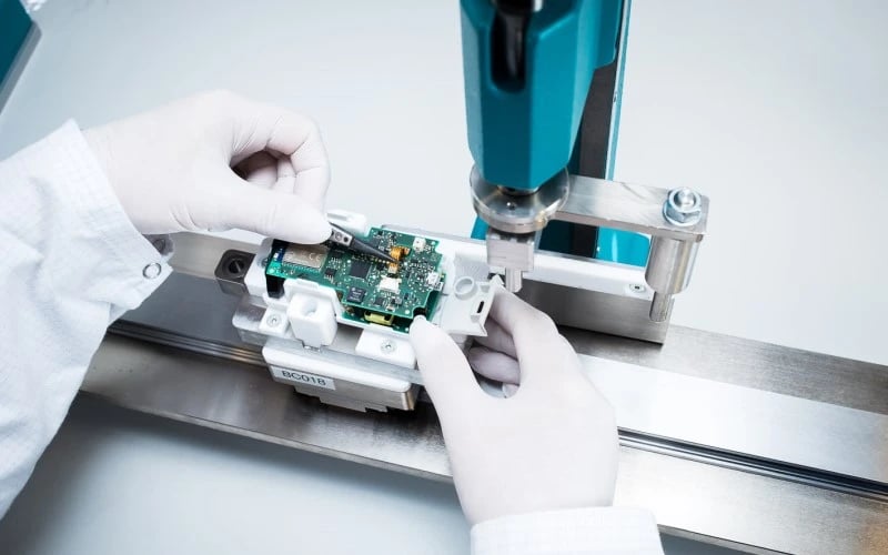 Two hands wearing white gloves placing an electronic component on a rectangular green printed circuit board mounted inside a small white plastic casing.  