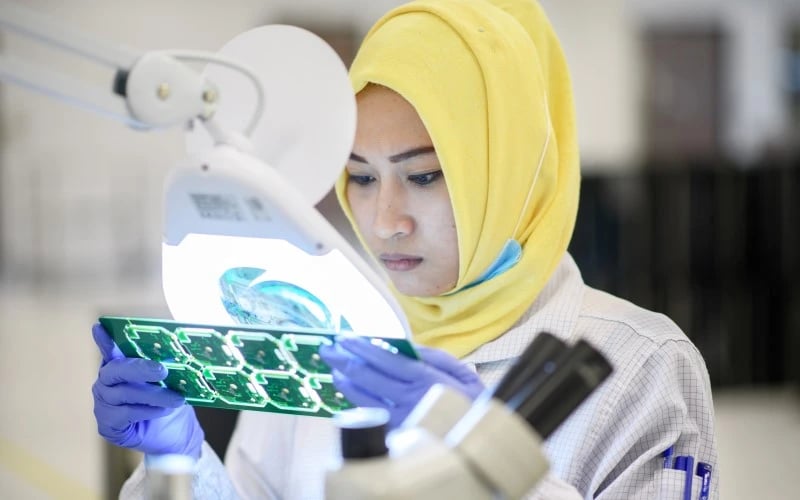 A female electronics operator wearing a yellow headscarf and a white protective jacket inspecting a panel of PCBs under an illuminated magnifying glass. 