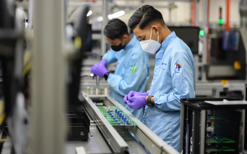 Zwei männliche Mitarbeiter in der Elektronikfertigung, die blaue ESD-Mäntel und lila Handschuhe tragen und elektronische Bauteile mit Durchgangsbohrungen auf einer Leiterplatte anbringen.
