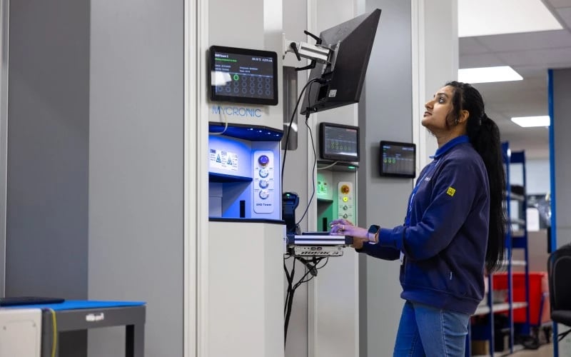 A female production operator looking at a display monitor in front of a Mycronic surface mount reel handling machine.  