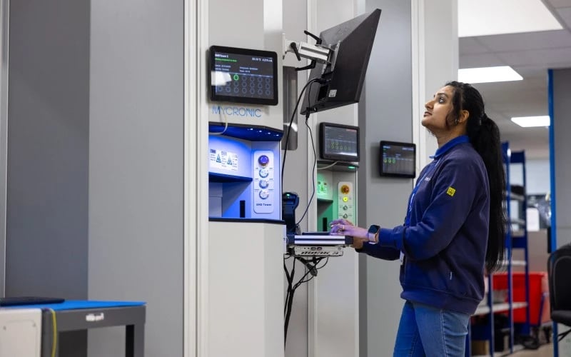 Eine Produktionsmitarbeiterin schaut auf einen Bildschirm vor einer Mycronic-Maschine für die Oberflächenmontage von Rollen.