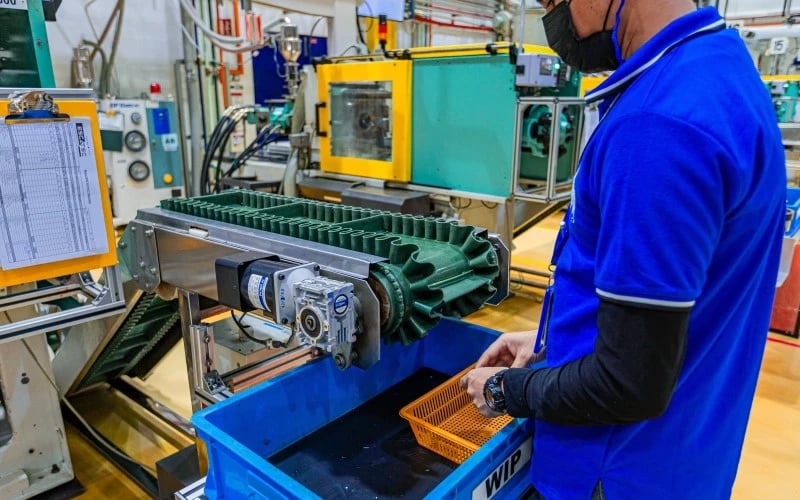 A production operative wearing a blue polo shirt working at the end of a plastic injection moulding machine waiting for the final plastic parts to come off the machine. 