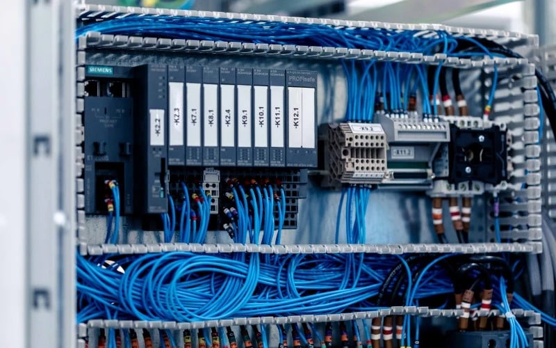 A control cabinet assembly with a large number of blue cables inside fitted to various electrical components like fuses, relays and circuit breakers within grey plastic trunking.