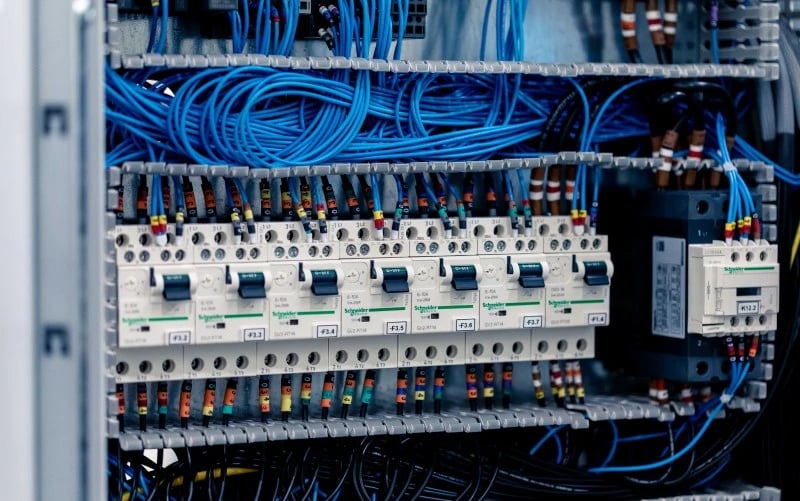 Eine Schaltschrankbaugruppe mit einer grossen Anzahl blauer Kabel im Inneren, die mit verschiedenen elektrischen Komponenten wie Sicherungen, Relais und Leistungsschaltern in grauen Kunststoffkanälen verbunden sind.