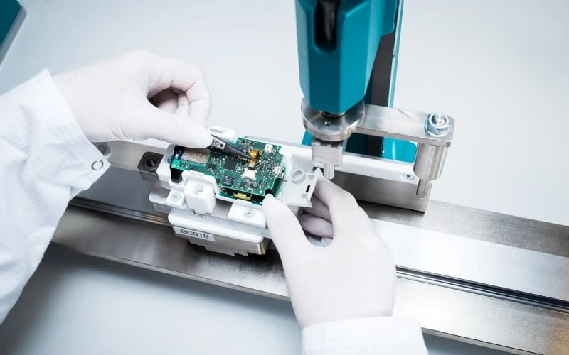 Two hands wearing white gloves placing an electronic component on a rectangular green printed circuit board mounted inside a small white plastic casing.  