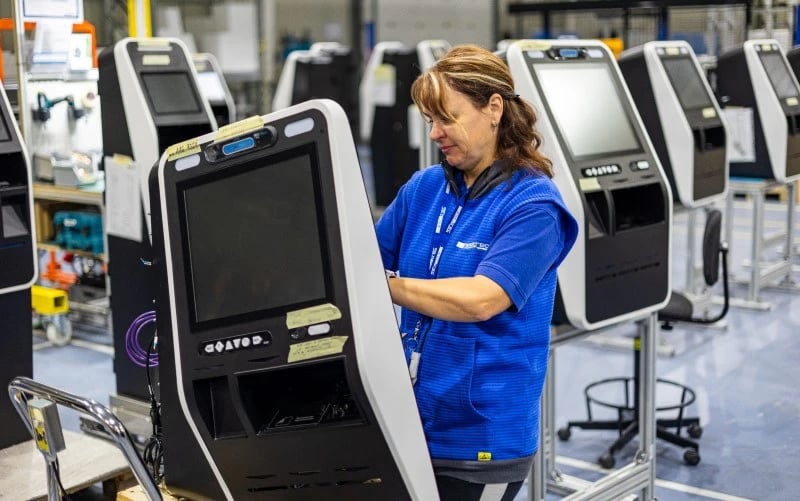 Eine Produktionsmitarbeiterin in blauer Kleidung arbeitet am Fliessband, umgeben von grossen schwarzen Kiosken mit Touchscreen-Displays.