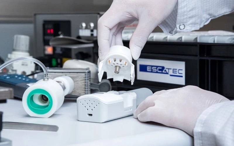 A close up of two hands wearing white gloves assembling part of a white hand-held medical device on a bench in front of test equipment and a black box with an ESCATEC sticker on it.