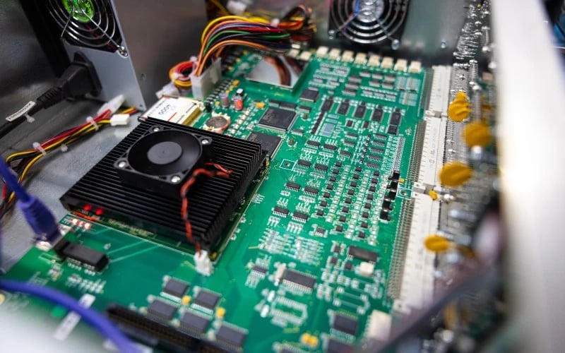 A close up of a green PCBA consisting of lots of semiconductors and wires along with a large black heatsink and fan assembly.  