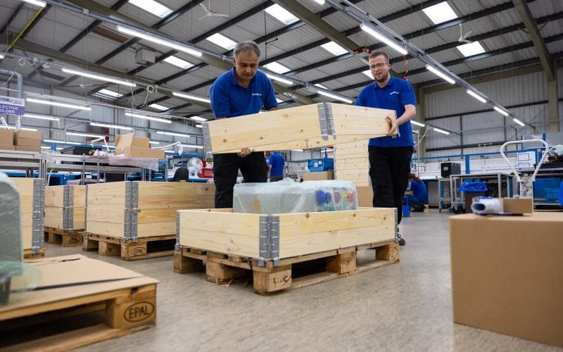 Two male production operatives working together holding the top half of a wooden packaging crate preparing to securely package a large and heavy electrical appliance sitting on a wooden pallet.