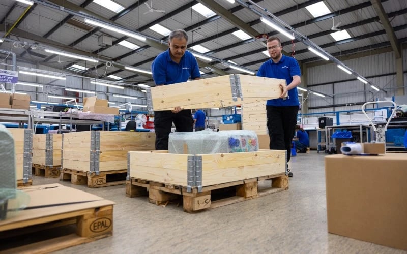 Zwei männliche Produktionsmitarbeiter arbeiten zusammen und halten die obere Hälfte einer hölzernen Verpackungskiste, um ein grosses und schweres Elektrogerät, das auf einer Holzpalette steht, sicher zu verpacken.  